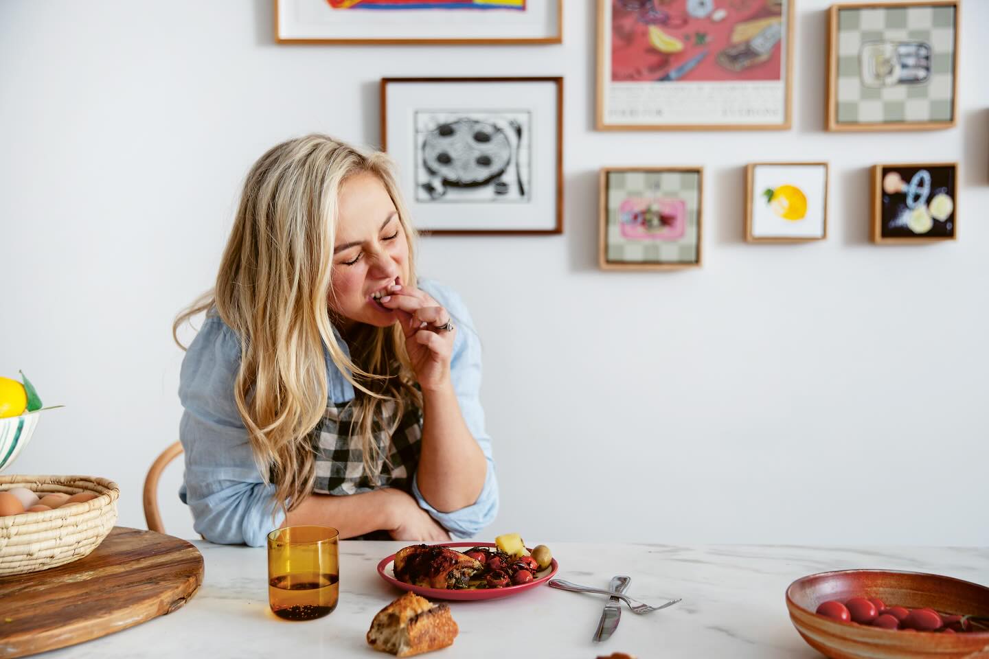 Woman eating at a table with various items on a white wall with framed pictures.
